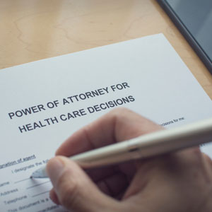 A hand holds a silver pen over a “POWER OF ATTORNEY FOR HEALTH CARE DECISIONS” document on a wooden surface